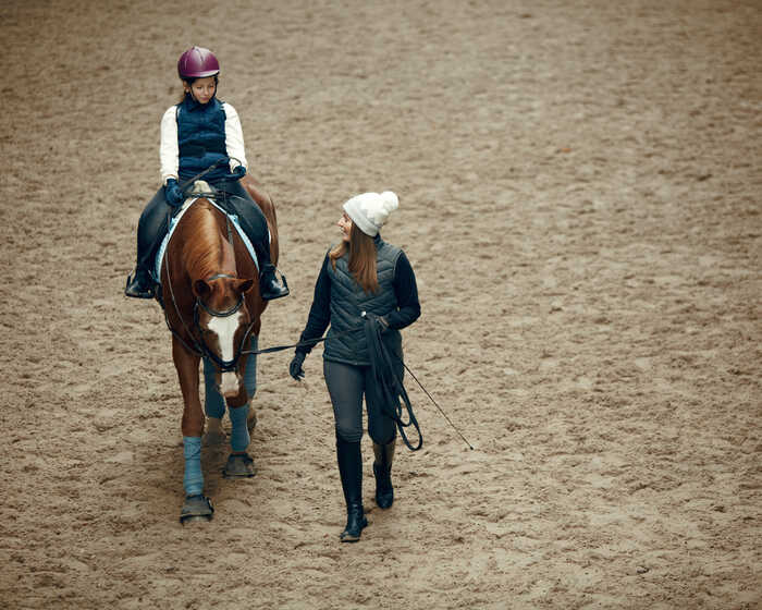 A child rides a horse while an adult walks alongside in an indoor arena.